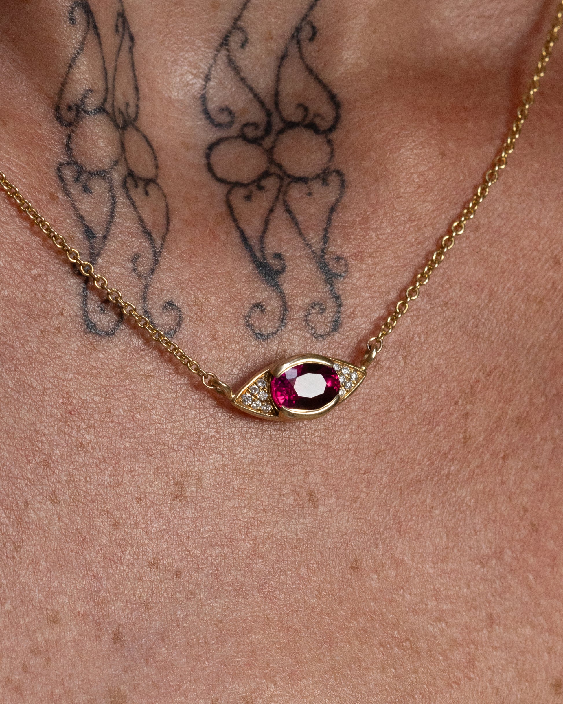 Close-up of the Pupil Pendant necklace, featuring a solid 14k gold eye-shaped pendant with a 1.7-carat oval-cut pink tourmaline and lab-grown white diamonds, worn on a person with visible butterfly tattoos on their chest. The rolo chain adds a delicate touch to the design.