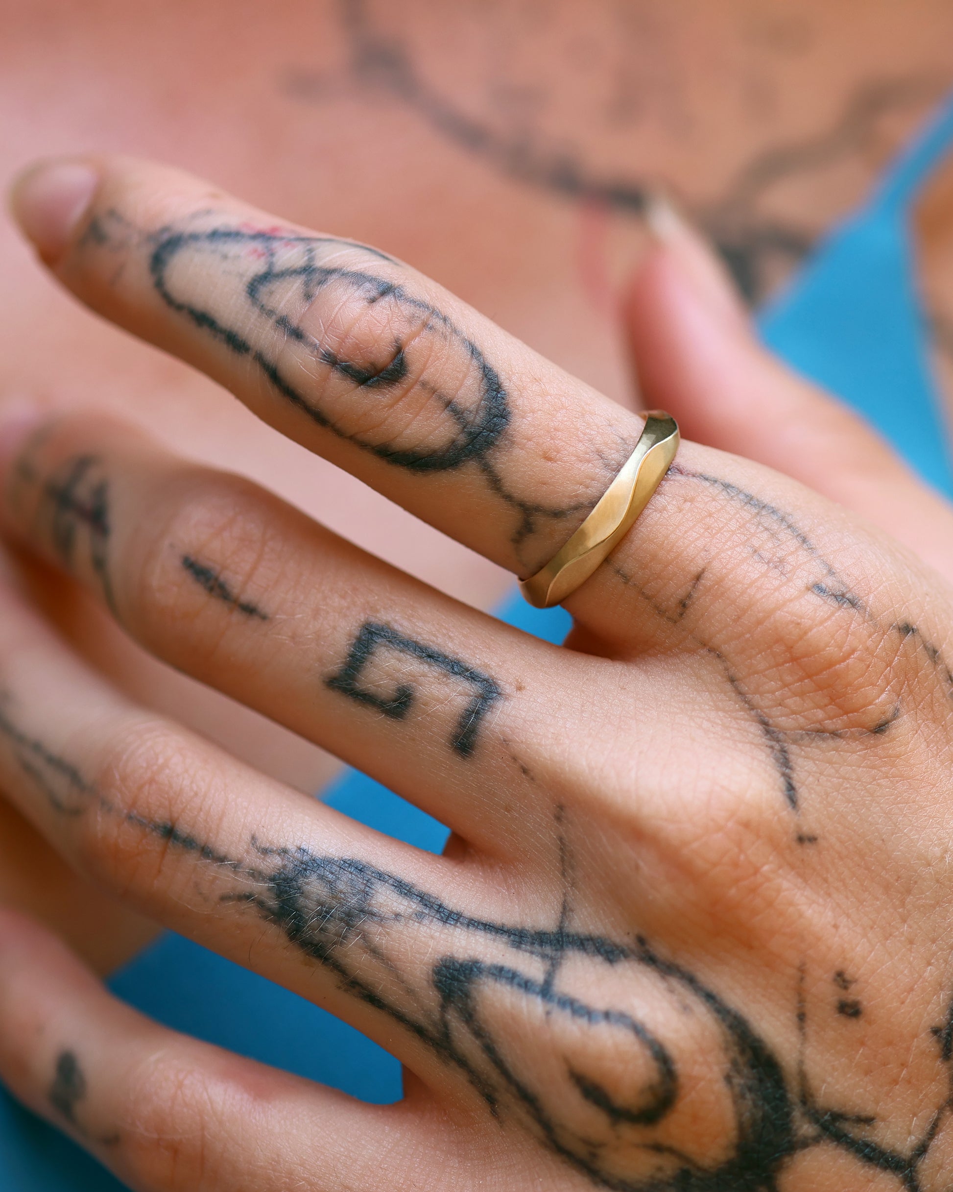 A hand adorned with tattoos wearing the Ribbon Ring, a solid 14k gold band with a subtle ruffled texture. The ring's elegant yet bold design complements the intricate tattoo artwork.