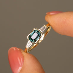 A close-up of a gold ring with a green emerald-cut center stone and baguette side stones, held between two fingers against a neutral gray background.