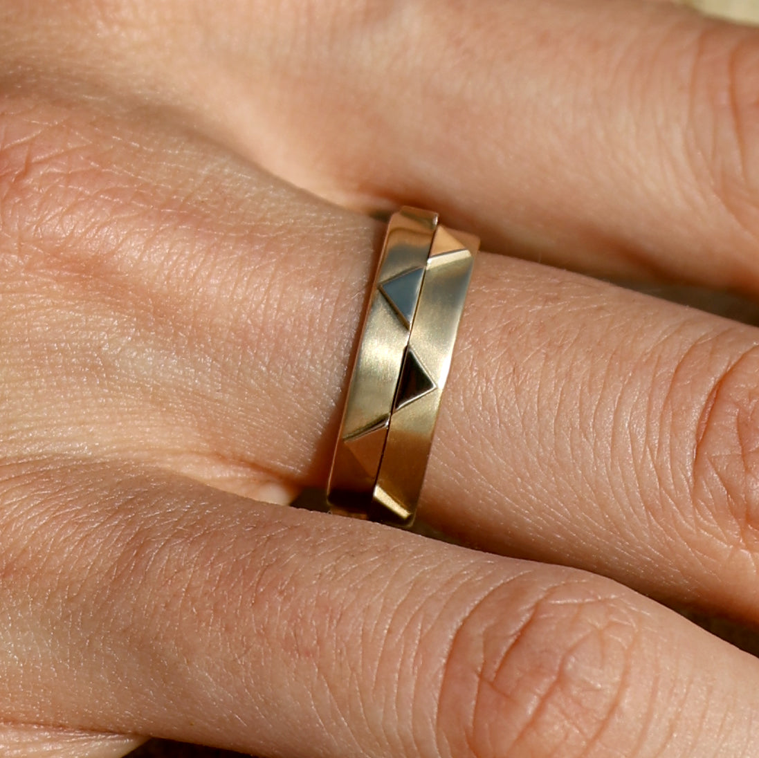 Close-up of two solid 14k gold geometric rings stacked together on a finger. The rings feature recessed triangular patterns and a mix of matte and polished finishes, creating a bold and modern look. 