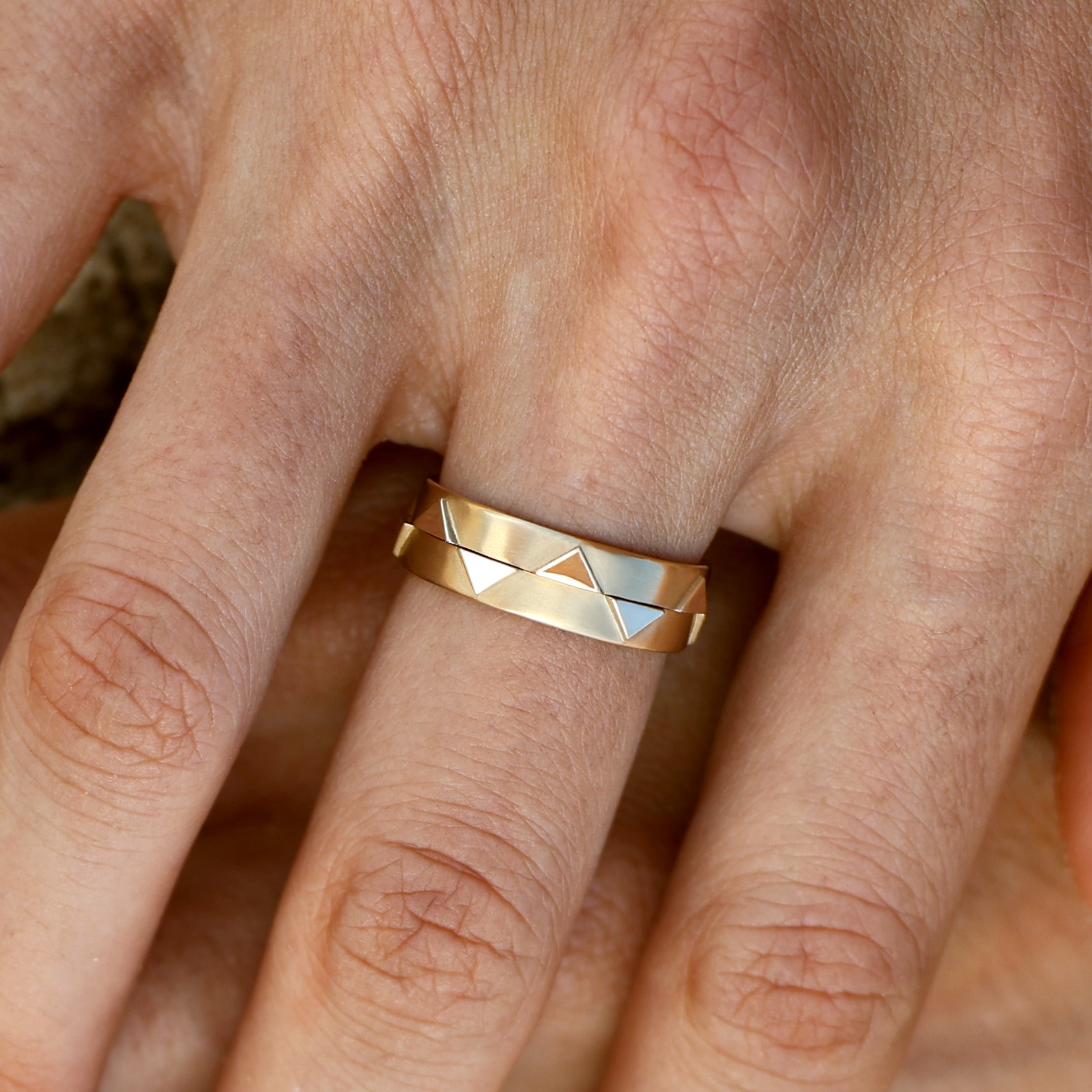 Close-up of a hand wearing two solid 14k gold geometric rings stacked together. The rings feature recessed triangular designs and a polished finish, showcasing a modern and versatile style.