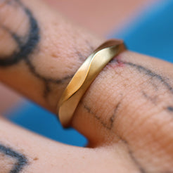 The Ribbon Ring, a solid 14k gold band with a subtle ruffled texture, worn on a tattooed finger. The matte finish highlights the ring's elegant and understated design. 