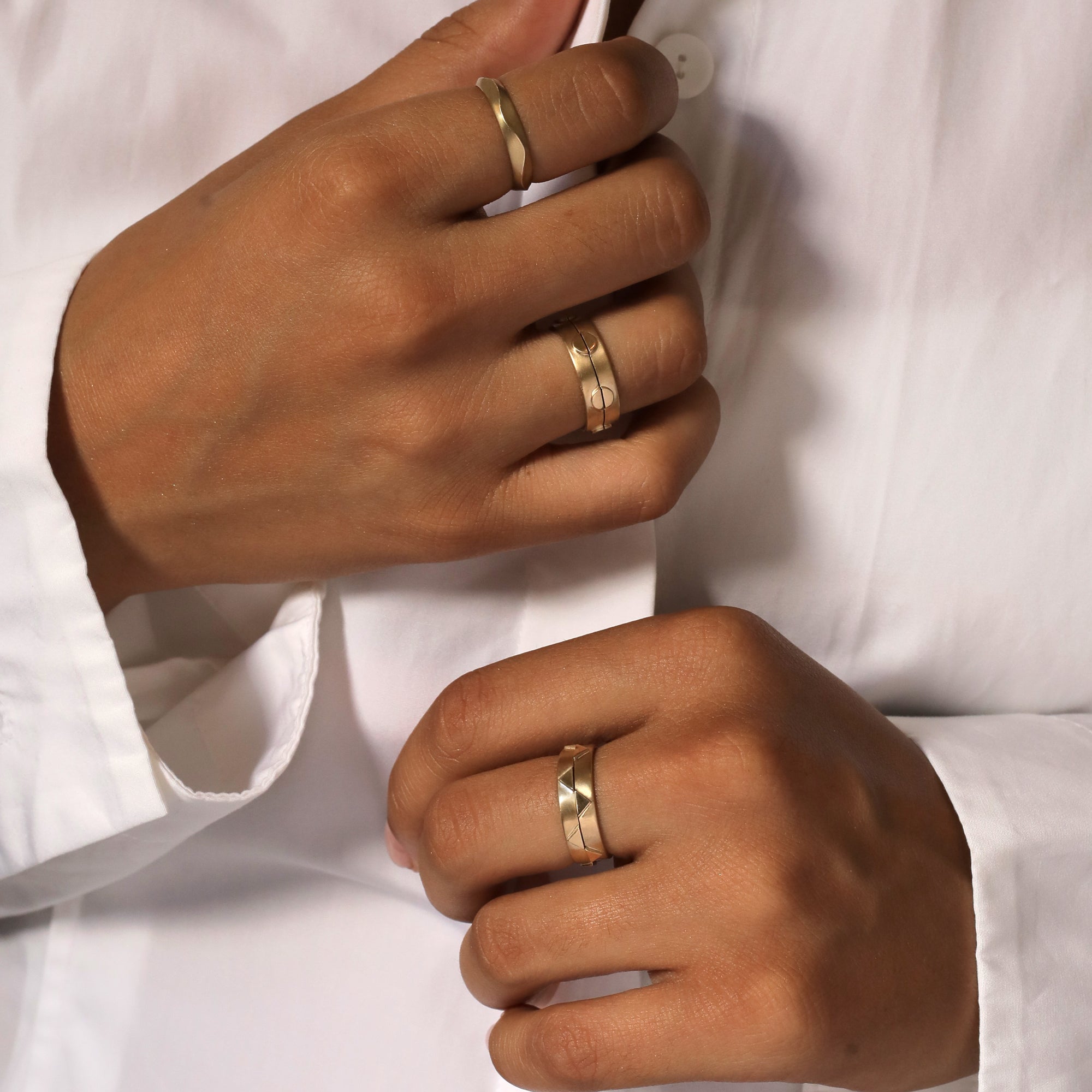 A person wearing three solid 14k gold rings with unique designs on both hands. The rings include a geometric band with triangular details, a sleek ribbon-style band, and a minimalist textured ring. The polished gold contrasts beautifully with a white shirt, creating an elegant look. 