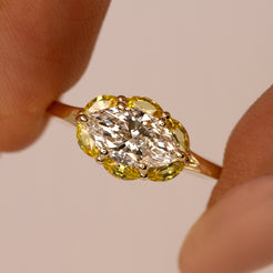 A close-up of a yellow gold ring held between fingers, featuring a marquise-cut clear center stone surrounded by a halo of yellow stones on a warm beige background.