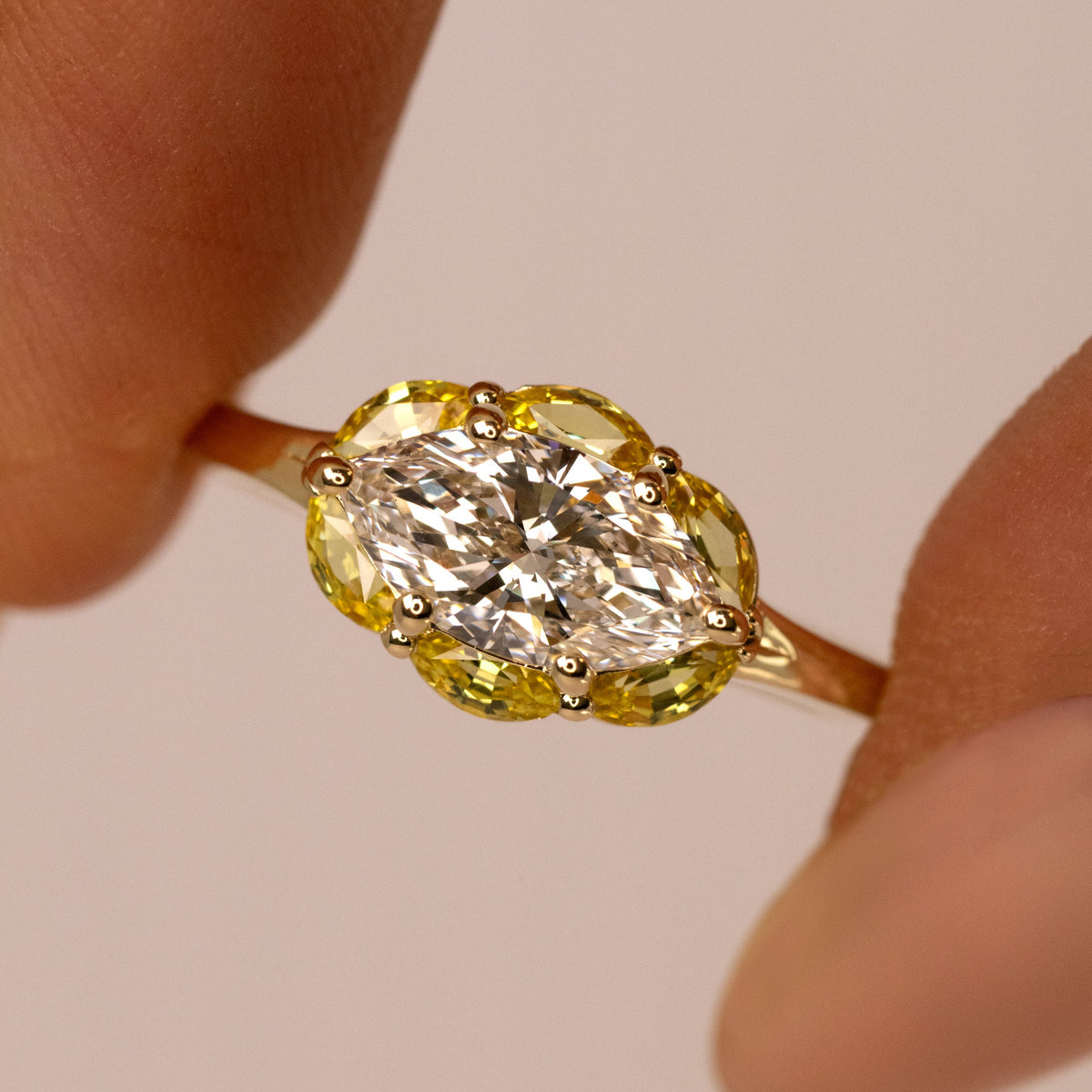 A close-up of a yellow gold ring held between fingers, featuring a marquise-cut clear center stone surrounded by a halo of yellow stones on a warm beige background.
